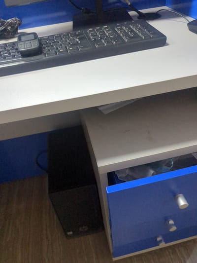 The image displays a close-up view of a desk area, likely in an office or home setting in Fagge A, Nigeria. The primary subjects are a black keyboard and a black computer tower. The keyboard is positioned on a white desk surface and has various buttons and keys visible. A small, black device with a screen and buttons, possibly a microphone or a small electronic device, is attached to the keyboard.
To the right of the desk, there is a blue cabinet with silver knobs. The cabinet appears to be part of a larger furniture unit. Behind the keyboard and desk, a portion of a white shelving unit is visible, with some white and possibly plastic items or bags inside. The background is a mix of these furniture elements and a textured surface that could be a carpet or flooring.
The lighting suggests it is daytime, but no specific time can be determined. There are no people visible in the image, and the overall scene is static, implying no particular activity is taking place at the moment the photo was taken. The image is taken from a slightly angled perspective, focusing on the details of the desk setup. No visible text is present in the image.