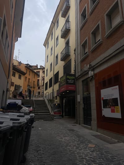 The image shows a narrow, cobblestone alleyway in Segovia, Spain, located at 40.947 latitude and -4.119 longitude. The alley is bordered by two tall buildings, both with windows and balconies. The left building is a pale yellow color, while the right building is a reddish-brown. There are several people walking in the alley, and some are sitting at tables on a small patio. The buildings are in a dilapidated state. On the right side of the alley is a storefront for a business called "Arcadia Salon de Juegos". The storefront has a red sign with the business name and a green sign that says "Poreto." There is a sign above the doorway that says "APUESTA." There is a smaller storefront next to it that is displaying a poster for a local business called "Bankinter." The image shows a view from the ground level of the alley, looking up towards the buildings. The alley appears to be in a state of disrepair. There are several trash bins on the left side of the alley. The sky is overcast.