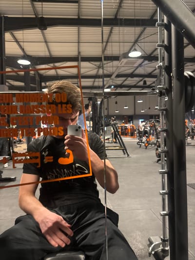 A selfie captured in a gym mirror reveals a person holding a phone, reflecting the camera. The gym's industrial ceiling with metal beams and overhead lighting is visible in the background. Weight training equipment, primarily in black and orange, is sparsely scattered throughout the gym. The person is wearing a black shirt and pants, and parts of a weight bench are reflected. The mirror has some orange text on it.