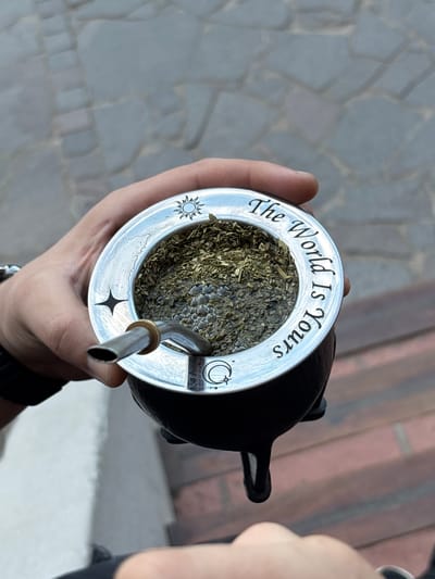A person holds a mate gourd filled with yerba mate and a bombilla (straw) in their hand. The gourd has a silver rim with the phrase "The World Is Yours" engraved around the edge, accompanied by small sun, star, and moon symbols. The background suggests an outdoor setting, with stone pavement visible. Based on the context of a mate gourd, the location may potentially be Villa General Belgrano, Argentina.