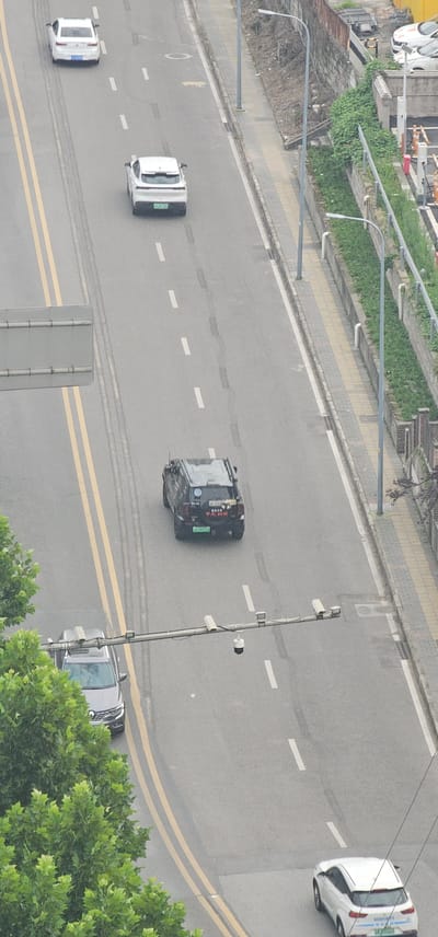 The image shows an aerial view of a street in Jiulongpo District, China. In the foreground, a silver SUV is parked on the left side of the road, partially obscured by lush green trees. A traffic camera gantry extends across the road, equipped with multiple cameras. Further down the road, a black SUV is driving in the right lane. The road has double yellow lines marking the lanes and white dashed lines indicating lane divisions. To the right of the road, a sidewalk runs alongside a retaining wall covered in vegetation, with lampposts and a fence. Further up the road, two white cars are visible, one in the left lane and another in the right lane. A white car is also seen at the bottom right of the frame. The scene depicts a typical urban street environment with moving and parked vehicles.