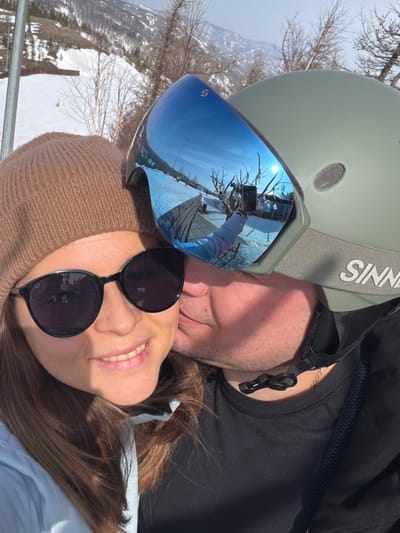 An image depicts two individuals in a close-up at a ski resort in Zelenaya Polyana, Russia. The female subject, positioned on the left, has long brown hair and wears a brown knit beanie, black sunglasses, and a light blue jacket. She is smiling towards the viewer. The male subject, positioned on the right, is shown kissing the female subject's left cheek. He wears a khaki green ski helmet with a "SINNER" logo and blue mirrored ski goggles, bearing "SCOTT" and "CONTRAST" branding. The goggles reflect a snowy ski slope, bare tree branches, a bright sun, a fence or barrier, and the individual taking the photograph (likely the female subject) holding a smartphone. The background reveals a snowy mountain slope with sparse bare trees and a portion of a ski lift structure under a bright, sunny sky.
