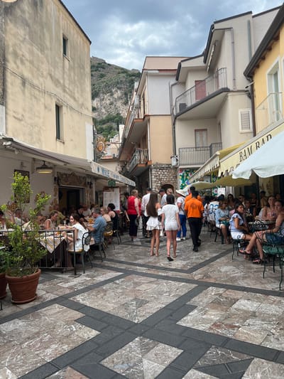 This image captures a bustling street scene in Taormina, Italy, likely in the historic center. The narrow street is lined with buildings, many of which appear to be restaurants or shops with outdoor seating. The architecture suggests a Mediterranean style, with stucco facades and balconies. People are seen dining at tables set on the cobblestone pavement, which is decorated with a marble pattern and dark grey borders.

In the foreground, a potted plant stands to the left, and the patterned pavement dominates the lower portion of the frame. Further back, several groups of people are walking and dining. On the left, a restaurant with a yellow and white awning, "Myle," is visible, with patrons seated at tables. To the right, another establishment with a yellow awning, possibly "BAM BAR," is also active, with people seated under umbrellas. The middle ground of the street is filled with pedestrians, many of whom are dressed in casual summer attire. In the background, a steep, rocky hillside with some vegetation and distant structures is visible under a cloudy sky. The overall atmosphere is one of a lively, tourist-oriented street in a popular Italian town.