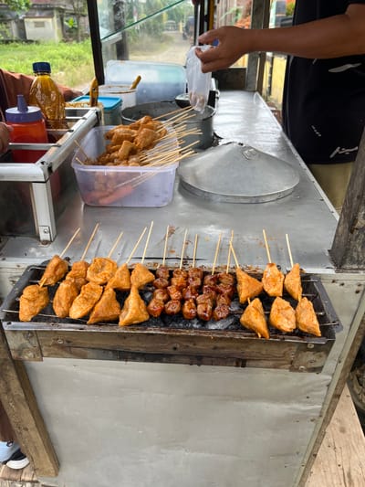 The scene depicts a street food vendor's cart in Matangpanyang, Indonesia, where various skewered items are being grilled and prepared outdoors during the daytime.
In the foreground, a metal charcoal grill contains multiple skewers of food. On the left side of the grill, approximately twelve skewers hold light orange-brown, irregularly shaped fried items, some triangular and some rounded, suggestive of fried tofu or fish cakes. On the right side, about ten skewers bear smaller, darker brown, glazed, spherical items, likely meatballs or similar protein skewers. Black charcoal embers are visible beneath the grill rack.
Above the grill, a metal cart forms the primary workspace. On the left side of the cart's metal countertop, a clear plastic bottle with a blue cap contains a thick, yellowish-brown liquid. Adjacent to it is a red squeeze bottle with a purple nozzle, presumably holding chili sauce. A clear plastic rectangular container holds numerous ungrilled or partially prepared skewered food items, similar to those on the grill, along with a few longer, reddish items (possibly sausages). Several other metal and plastic containers, some with lids (one light blue, one white), are arranged on the countertop.
On the right side of the countertop, a large, conical metal lid rests, possibly covering a pot or steamer. A person, visible from the mid-torso to the upper arm, wearing a black t-shirt with white patterns, is holding a small, clear plastic bag containing food items over a metal bucket or container. Another person's arm, dressed in a brown sleeve, is partially visible on the far left, near the sauce bottles. Their lower leg and a black shoe with a white sole, worn with a blue and white striped sock, are also visible at the bottom left.
The background, out of focus, shows blurred greenery consistent with trees and grass, along with indistinct structures or buildings, indicating an outdoor or roadside setting. The ground beneath the cart is a light-colored surface, possibly concrete, with some wooden planks or supports visible at the edges.