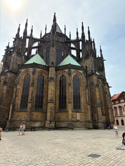 The image shows the exterior of a large Gothic cathedral, likely St. Vitus Cathedral in Prague, Czechia. The cathedral is constructed of weathered stone and features intricate architectural details, including spires, buttresses, and arched windows. Two green-roofed sections of the cathedral are visible. The cathedral is situated on a cobblestone plaza, where several people are walking. The sky is cloudy, suggesting it is either overcast or early morning/late afternoon. The overall scene conveys a sense of historical grandeur and a typical day for tourists visiting a famous landmark.