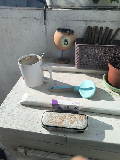 An outdoor still life scene is captured on a weathered, white wooden surface, likely a balcony railing or table, in Rivne, Ukraine. Sunlight casts distinct shadows, indicating a bright day.
Dominating the foreground is a white ceramic mug, stained with a brown residue, suggesting it recently held coffee or tea. A thin, metallic spoon rests inside. To the right of the mug, a light blue and beige brush lies flat. In front of the brush, a small rectangular box with purple and white text, labeled "DELIA PURPLE CAPSULE," is positioned. Beneath the "DELIA" box is a cream-colored case with an embossed, intricate pattern, likely for glasses or a similar item.
In the background, a basket filled with wooden clothespins is visible. To the left of the basket, a small, round, amber-colored object with the number "5" printed on it sits atop a dark base, resembling an ashtray. A terracotta pot containing dark soil and a green plant is situated to the right of the basket. The overall environment suggests a domestic setting, possibly a balcony or patio, with elements of everyday use. The background shows a blurred, light-colored concrete wall and a glimpse of an outdoor setting.