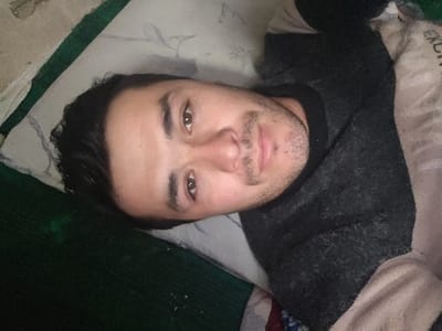 A man with dark hair and a light complexion is lying down. He is wearing a dark sweater with a light-colored edge. He has stubble on his face and is looking directly at the camera. He is on a pillow with a white and blue pattern. The background consists of a dark green and beige fabric.