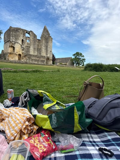 The image shows a picnic setup in front of the ruins of Minster Lovell Hall in West Oxfordshire, UK. A blue and white checkered blanket serves as the picnic base, upon which are arranged: a grey backpack, a beige handbag, a green and yellow shopping bag, a package of "Hoops" snacks, a yellow and white checkered cloth, a partially visible clear plastic container with food, and a can of beer. The ruins of Minster Lovell Hall are visible in the background, along with a green lawn and a sky with scattered clouds.