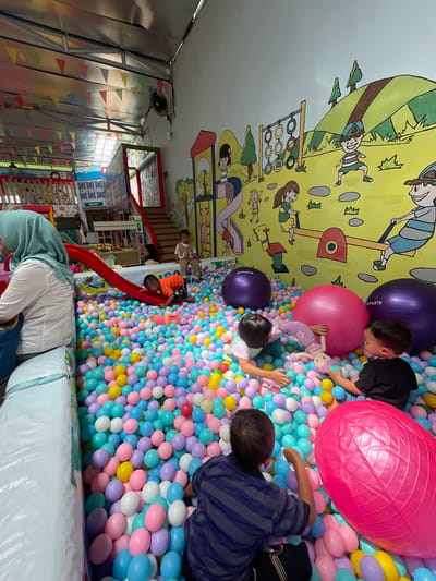 The image captures an indoor playground, possibly in Sinduagung, Indonesia. The dominant feature is a large ball pit filled with pastel-colored plastic balls, with several children playing inside. Three large exercise balls (purple and pink) are also within the ball pit. The wall behind the pit is decorated with a mural depicting children playing on playground equipment. A red slide leads into the ball pit from a raised platform, where a few other children are visible. A woman wearing a headscarf stands near the edge of the ball pit, possibly supervising the children. The left side of the image shows a play area with a multi-level structure and colorful decorations. The environment appears to be a commercial indoor playground.