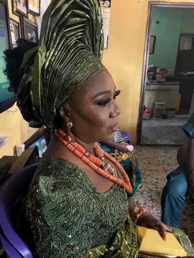 A mature, dark-skinned woman wearing an elaborate olive green, pleated head wrap and a matching green beaded and sequined dress is seated indoors. She has a coral necklace and bracelet. A person in a teal-colored garment is partially visible above her. The scene includes a mirror reflecting a cluttered room, framed pictures, and various objects on a table. The location is Warri, Nigeria.
