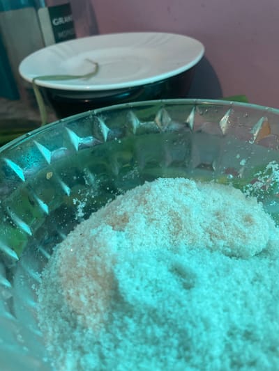 A close-up, slightly blurry, high-angle shot reveals a clear glass bowl containing a large, compacted mass of white granular material, possibly salt or sugar. The bowl features a textured exterior with diamond-shaped indentations. To the left, partially obscured, are a white plate stacked on a black object, potentially a container or another plate. The background is indeterminate, showing hints of green and pink. It is impossible to determine if this image was taken in Olginka, Russia.