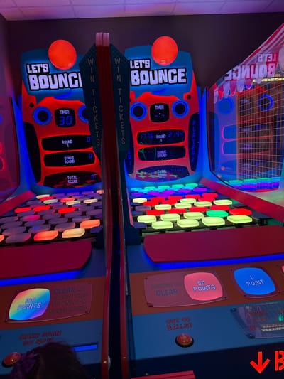 The image shows a "Let's Bounce" arcade game, potentially at an entertainment center in Columbus, Ohio, USA. The game is designed for two players and involves bouncing balls to hit lit-up tiles. The display shows a timer, round number, bonus round status, and total score for each player. Instructions on the machine explain the point system and how to earn bonus rounds. An orange ball is present on one side of the game.