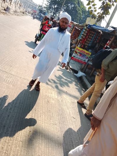 The image depicts a street scene in Shibpur, Bangladesh, under bright daylight. In the foreground, slightly left of center, a man with a dark beard and a serious expression stands on a paved surface. He wears a white turban, a long white tunic (thobe/jubbah), and brown sandals. His shadow extends diagonally towards the lower left.
To the immediate right of this man, another individual is partially visible, wearing khaki pants and dark sandals, with a dark bag or garment draped over their shoulder/back. In the lower right corner, the lower portion of a third person wearing a light-colored traditional garment is visible.
In the mid-ground, behind the central figure, a green and red auto-rickshaw, possibly an E-rickshaw, is parked or moving slowly. A red sign with "FOR RENT" in white English text is visible on its side. Other individuals are partially visible inside or near the auto-rickshaw.
To the right of the street, along a curb, are roadside stalls. One stall is visible with numerous packaged snacks and goods hanging for sale. A yellow signboard with Bengali text, reading "উল্লাসি প্রিমিয়াম কর্ন" (Ullashi Premium Corn) among other details, is prominent next to the stall. The pavement surface shows distinct parallel grooves.
The background features additional buildings and greenery, suggesting a busy urban or semi-urban environment. The lighting indicates direct sunlight, casting sharp shadows from people and objects onto the pavement.