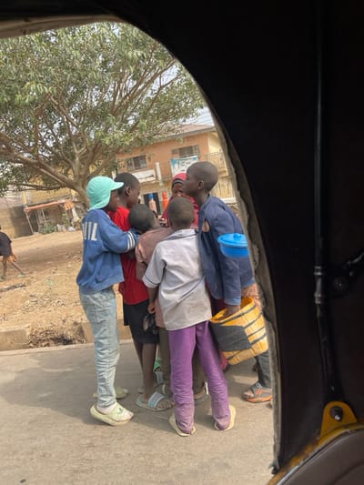 The image, framed from within a vehicle, depicts an outdoor scene in Samaru, Nigeria, during daylight hours. A group of seven children are clustered on a paved surface.
In the foreground, a child with their back mostly to the viewer wears a light grey shirt, purple trousers, and light-colored slip-on shoes. To their left, a child in a light blue cap, blue long-sleeved shirt, and light blue jeans stands, with one arm around another child wearing a red shirt. Partially obscured among the cluster is a female child wearing a reddish head covering. To the right of the central cluster, another child in a dark blue jacket holds a yellow plastic jerrycan or bucket with black strapping in their left hand, and a blue lidded bowl is hooked onto their left shoulder. This child also wears dark trousers and dark sandals. Other children are partially visible within the huddle, wearing various tops and shorts/trousers. The children appear to be interacting or observing something within their immediate group. Footwear varies from sandals to slip-on shoes.
To the left of the paved area is an unpaved dirt ground with some sparse vegetation. In the mid-ground, a large deciduous tree with mostly bare branches is prominent. In the background, a two-story building with a light brown/orange facade, featuring multiple windows and a balcony with a dark railing, is visible. Adjacent structures are partially seen. The sky is clear, suggesting fair weather. Another lone figure is partially visible on the dirt path in the far left background.