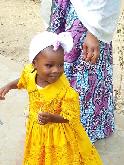 In Garun Gawa, Nigeria, a young girl in a yellow dress stands holding a lollipop. She wears a white head covering with a pink bow. To her right stands an adult woman in a colorful printed dress with a white top. They are on a paved surface near a dirt area. The girl is looking to the side.