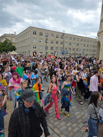 A crowded street scene in Warsaw, Poland, features a large group of people participating in a Pride parade. The participants, of various ages and backgrounds, are walking down a cobblestone street lined with multi-story buildings. Many individuals are adorned in colorful clothing, rainbow flags, and accessories, signifying their support for the LGBTQ+ community. Some are holding flags, banners, and what appear to be brochures. The sky above is overcast with clouds. In the background, a large, light-colored building with many windows dominates the cityscape. The presence of streetlights and architectural details suggests an urban setting. The atmosphere appears festive and celebratory, with people engaging with each other and moving in the same direction.