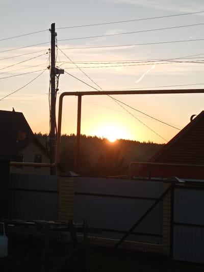 A low-angle, medium shot captures a sunset scene with a prominent utility pole and overhead wires in the foreground, positioned left of center. The sky is a gradient of pale blue and soft orange, with a faint contrail visible near the top right. A line of dark trees forms a silhouette against the bright horizon. In the middle ground, partially obscuring the trees and sky, are rooftops of houses and a metal fence. The fence, made of grey corrugated metal panels with brick pillars, runs horizontally across the frame. To the left, the dark roof and side of a house are visible. A yellow gas pipe runs horizontally across the upper middle of the image, supported by vertical structures. In the lower foreground, a dark wooden structure, possibly a platform or makeshift table, is visible on the ground, with sparse vegetation. The overall impression is of a rural or suburban setting at dusk. This image was taken in Degtyarsk, Russia.