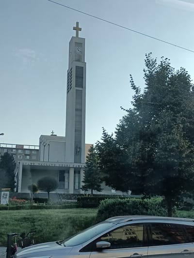 This media file is a photograph taken outdoors on a clear day, likely in the late afternoon given the lighting. The scene features a modern church with a tall, rectangular bell tower topped with a cross. The tower has a clock face and vertical bands of windows. Below the tower, the church has a wider structure with a covered entrance area, displaying the text "SVATY VACLAVE, NEDEJ ZAHYN" in large, raised letters.
In the foreground, a silver car is partially visible, reflecting the surrounding buildings and trees. A bicycle is parked to the left of the car. Behind the church, a large, leafy tree dominates the right side of the frame, its branches casting shadows. In the background, a multi-story apartment building can be seen. The area in front of the church is landscaped with grass, bushes, and some flowering plants. The overall impression is of an urban or suburban setting in Prague, Czechia. No people are clearly visible in the frame.