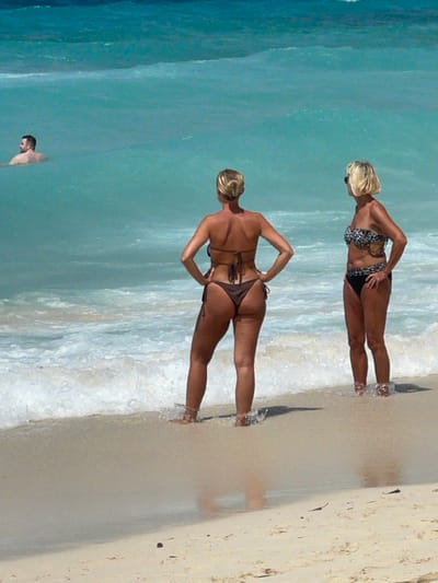 The image displays a beach scene in Zanzibar, Tanzania, with three individuals. In the immediate foreground, two adult females stand at the waterline on light-colored sand. The woman on the left, with blonde hair styled in a bun, is oriented with her back to the viewer, wearing a dark brown string bikini and posing with her hands on her hips. Her lower legs and feet are partially submerged in the shallow, white-foamed surf. To her right, a second woman with short blonde hair and sunglasses stands facing slightly left, wearing a black and white leopard print bikini top and black bikini bottoms. Her lower legs are also immersed in the breaking waves. In the background, further out in the clear, light-turquoise ocean, an adult male is visible from the chest up, facing rightward. The water shows gentle ripples and white foam where waves break on the shore.
