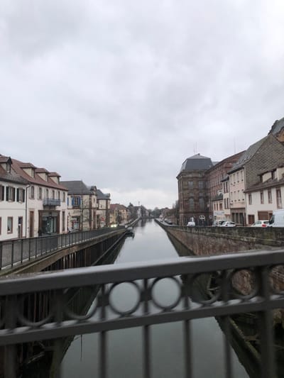 The image depicts a tranquil canal scene in what appears to be a European town, captured on an overcast day. The perspective suggests the photo was taken from a slightly elevated position, possibly from a bridge or an upper floor, looking along the canal.

The top portion of the image is dominated by a pale, cloudy sky, indicating a dull or wintery day without direct sunlight. The lighting is soft and diffused.

Running horizontally through the midground is a calm, dark body of water, which is likely a canal or a narrow river. Its surface reflects the muted tones of the sky and the surrounding buildings, creating a smooth, almost glassy appearance.

On both sides of the canal, rows of multi-story buildings line the banks.
On the left bank (from the viewer's perspective), the buildings display a mix of architectural styles. Some have steeply pitched, tiled roofs, while others feature flatter, darker roofs. Facades are painted in shades of white, beige, and light brown, with visible windows, some of which have closed shutters, including a building with distinctive blue shutters. An external wooden or metal structure, possibly a walkway or a series of steps, runs along the water's edge in front of some buildings. A few bare-branched trees are scattered along this bank, reinforcing the impression of a colder season.

On the right bank, the buildings appear to be slightly grander in scale. One prominent building in the mid-distance features a dark mansard roof and a lighter-colored stone or stucco facade with numerous rectangular windows. Further down this bank, other buildings with varying rooflines and fenestrations are visible. Several cars, including a white van, are parked along the street that runs parallel to the canal on this side.

In the immediate foreground, on the right side of the image, a dark metal railing is prominently featured. It has a decorative pattern of circular elements connected by horizontal bars, partially obscuring the view of the canal and the buildings on the right bank. This railing suggests the presence of a bridge, balcony, or a waterfront promenade where the photo was taken.

The overall atmosphere is quiet and still, with no human activity or boats visible on the water. The architecture suggests a historical European setting. No discernible text is visible on any of the buildings or signs.