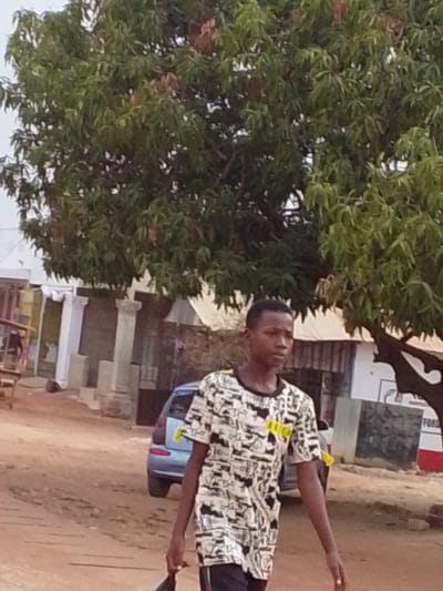 This full-body, outdoor shot captures a young person walking on an unpaved road in Dungu, Ghana, during what appears to be an overcast day or late afternoon, given the soft, diffused lighting and lack of harsh shadows. The subject is a male with dark skin, wearing a short-sleeved t-shirt featuring a complex black and white graphic pattern. The t-shirt has black sleeves and a black collar, and notable details include several small yellow rectangular patches on the t-shirt. One patch on the chest has partially visible text that reads "ROAD," while another near the right shoulder is less clear but might include "KEEP WA" or "NEW WA." He is also wearing dark trousers. He holds a dark object or bag in his left hand, and his gaze is directed slightly to his right, with a neutral expression.

Dominating the upper portion of the frame is a large, dense tree with plentiful green leaves, possibly a mango tree, providing significant shade. In the background, along the dirt road, there are various buildings and structures. These include constructions with corrugated metal roofs and distinctive white pillars or columns. A light blue car is partially visible, parked on the reddish-brown dirt road behind the person. The overall scene suggests a candid moment in a semi-urban or rural setting.