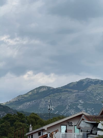 The image captures a view of the sky and mountains in Mrčevac, Montenegro. The sky is filled with overcast, gray clouds. In the foreground, the roof of a building with red tiles is visible, along with parts of its structure, including a chimney. To the left of the building, there are dense green trees. In the middle ground, a cell tower stands tall amidst the trees, with mountains stretching into the background under the cloudy sky.