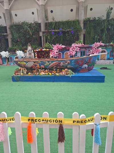 The image shows a brightly decorated, large-scale model of a boat, positioned outdoors on a green turf surface. The boat is adorned with a colorful mural depicting marine life, including turtles, fish, coral, and an anchor. It is overflowing with artificial flowers and fruit, creating a vibrant, festive display. Behind the boat, a lush green vertical garden is visible on a wall, with buildings that appear to be decorated with colorful facades and flowering trees. A white picket fence with yellow caution tape reading "PRECAUCIÓN" stretches across the foreground, with colorful tassels hanging from the fence posts. The overall scene suggests an outdoor public space or an event in Pampatar, Venezuela, designed for visual appeal and possibly as a decorative installation.