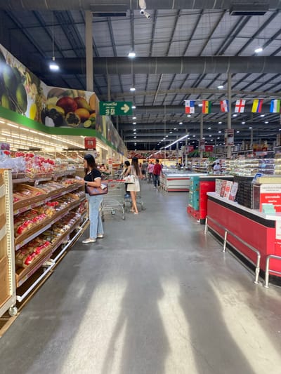 The image depicts an interior view of a large supermarket, possibly a hypermarket or warehouse store. The perspective is wide and long, showing a central aisle lined with shelves of merchandise. The setting appears to be brightly lit, likely with artificial lighting, indicating it's either evening or a windowless interior. The floor is a gray concrete, with some minor stains and cracks.
In the foreground, a woman wearing a black top and jeans is seen from the side, pushing a shopping cart. She is looking down at the shelves, which are filled with packaged food items, possibly baked goods or snacks, in clear plastic packaging with red accents. Further down the aisle, more shoppers with shopping carts are visible, though their details are less clear due to the distance and angle.
The shelves on the left side of the aisle are stocked with various products, with some promotional posters or images, including one featuring a vibrant display of fruits like mangoes, passionfruit, and pineapples. Above the shelves, rows of flags are hanging from the ceiling, suggesting a possible international theme or a display related to a specific event. Some of these flags are recognizable, including what appears to be the flag of Panama and the flag of Ghana, among others. There is also a green directional sign with Thai text and an arrow pointing upwards.
On the right side of the aisle, a prominent red display fixture is visible, likely a checkout counter or a promotional display. Shadows cast across the floor suggest the direction of the lighting. The overall atmosphere is that of a typical busy retail environment.
The location context of Baan Bo Phut, Thailand, suggests that the text on the signs and the types of products displayed might be consistent with a Thai market. The flags could represent a variety of countries, perhaps signifying the store's international offerings or a global event. No specific time of day or weather conditions are discernible from the interior scene. The emotions of the shoppers are not evident.