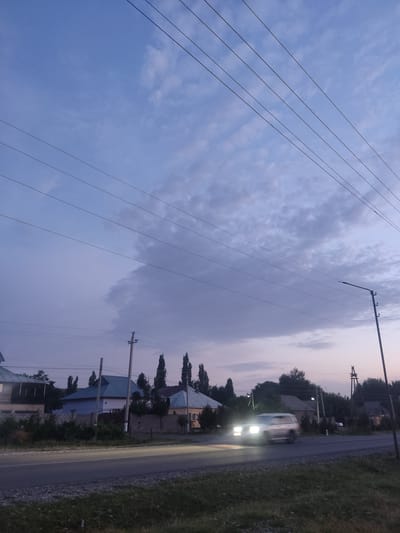 The image depicts a rural scene in Zertas Rural District, Kazakhstan, during dusk. A road runs alongside a grassy area, with houses and trees lining the opposite side of the road. Several utility poles with wires are visible, contributing to the rural aesthetic. A white van is driving on the road, its headlights illuminated, creating a bright streak of light. The sky is a gradient of blue and purple, with scattered clouds, suggesting the transition from day to night. The overall atmosphere is calm and serene, characteristic of a rural evening.