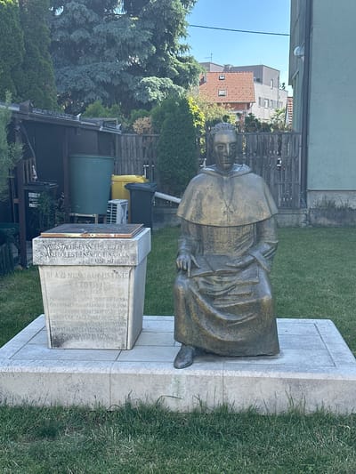 Here's a detailed description of the image:
The image features a bronze statue of Blessed Augustin Kazotic, a Croatian Dominican friar who was the Bishop of Zagreb in the 13th and 14th centuries. The statue depicts him in a seated position, wearing a bishop's robes and head covering. The statue is mounted on a stone base with inscriptions. The inscription on the monument states that he was "the most distinguished of Croatian people."
The statue is located outdoors, with a building and trees in the background. The statue is standing on a stone base sitting on the ground in a grassy yard. A large olive-green rain barrel and a pair of refuse containers are in the background. The sky is clear and blue. The location of the statue is likely in the city of Zagreb, Croatia, though there are no visual elements in the image that confirm the exact location within Zagreb.