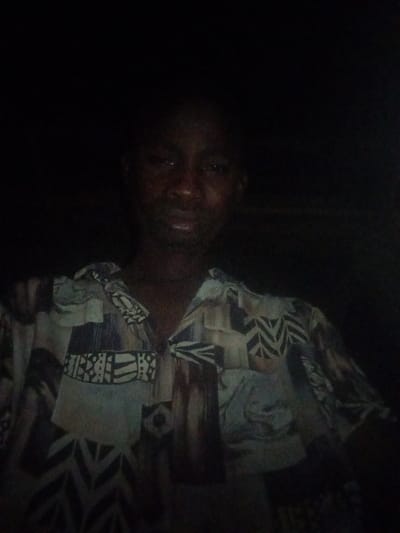 A dimly lit, frontal photograph depicts a dark-skinned individual from the upper torso up, centered against an indistinguishable dark background. The individual wears a short-sleeved, collared shirt featuring a complex pattern of geometric and abstract shapes in varying shades of beige, brown, blue, and black. The individual's eyes are directed towards the camera. No other subjects, objects, or environmental details are discernible within the frame. The scene is located in the city of Adonkolo, Nigeria.