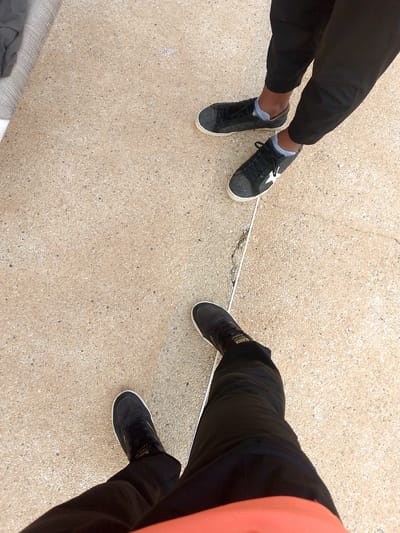 The image shows the lower bodies and feet of two individuals standing on a concrete surface. The concrete appears to be textured with small stones. A thin, white line or tape runs diagonally across the frame.
The person on the left is wearing black pants and black sneakers with white soles. One shoe has a white star emblem. They are also wearing light blue socks.
The person on the right is wearing black pants and black sneakers with white soles. The sneakers have a subtle star pattern. They are also wearing light blue socks.
The environment appears to be outdoors, possibly a sidewalk or public area. No specific city landmarks or distinctive features of Pedro González, Venezuela are visible.