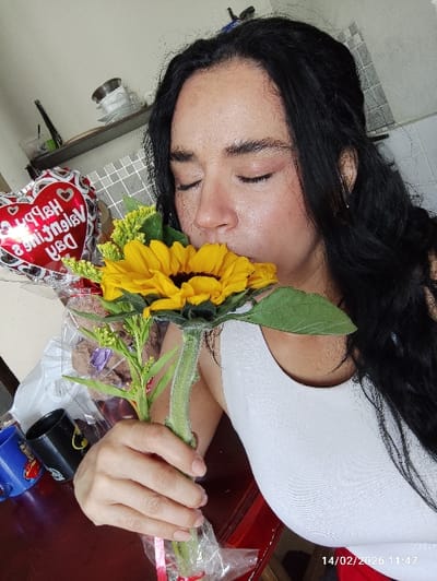 An overhead photograph captures a person embracing a bouquet primarily featuring a vibrant sunflower, appearing to smell or gently kiss the flower. The person is an adult with long, dark, wavy or curly hair that cascades around their head. They are wearing a plain white tank top or sleeveless shirt. Their eyes are closed, and their lips are slightly puckered, positioned directly against the dark center of the large yellow sunflower. One hand, with neatly kept, unpainted fingernails, holds the thick, fuzzy stem of the bouquet. The stems are wrapped in clear plastic, possibly with a hint of red ribbon.

The bouquet consists mainly of the prominent yellow sunflower, accompanied by smaller clusters of green-yellow filler flowers or buds and some green leaves.

The setting appears to be indoors. In the background, on a dark wooden surface (likely a table), a blue mug with a design and a black mug are visible. Behind these, a white plastic bag and another transparent plastic bag containing some brown, wrapped items (possibly a treat or small gift) can be seen. To the right of the person's head, a red heart-shaped foil balloon is partially visible, appearing slightly deflated or creased. The balloon features white text, which, when deciphered despite being upside down and mirrored, reads "Happy Valentine's Day" surrounded by smaller hearts. Below the balloon, a gray tiled surface, resembling a floor or countertop, is visible.

A digital date and time stamp, "14/02/2020 11:47," is present in the bottom left corner of the image, indicating the photograph was taken on Valentine's Day, February 14, 2020, at 11:47 AM. The overall impression is one of gentle affection and enjoyment, likely celebrating Valentine's Day.