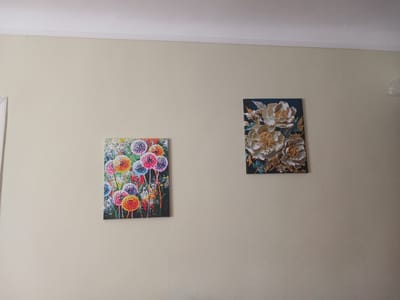 The image shows two abstract paintings hanging on a plain, cream-colored wall. The painting on the left features a vibrant bouquet of what appear to be dandelions in various colors, including pink, yellow, blue, and orange, against a dark background. The painting on the right displays a close-up of three large, intricately detailed flowers in shades of white and gold, set against a dark blue background. Both paintings are rectangular and hung at different heights. The ceiling molding is visible at the top of the image. The scene is devoid of any people or animals, and the overall environment is simple and uncluttered, suggesting an indoor setting. The city of Mamaivtsi, Ukraine, is not visually represented in this image.