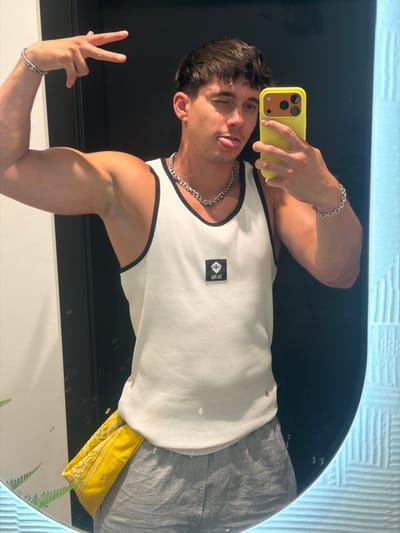 A young man is taking a mirror selfie in what appears to be a bathroom. He is wearing a white ribbed tank top with black trim, grey shorts, and a yellow bandana tucked into his waistband. He has a silver chain necklace and a silver bracelet on his left arm. The man is winking and sticking out his tongue, with his right hand holding up a peace sign. He is also holding a yellow phone in his left hand, which is angled towards the mirror. The background is a dark, possibly black, wall and a tiled surface that is white or light blue. There are some faint reflections in the mirror. The scene does not show any elements that would indicate the city of Alberobello, Italy.