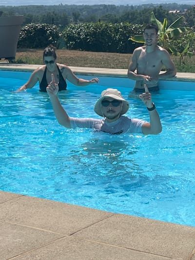 In Saint-Galmier, France, three individuals are present in a swimming pool on a sunny day. The person in the foreground, wearing a bucket hat, sunglasses, and a white t-shirt, is partially submerged and raising both index fingers. To their left, a woman in a black bikini is also in the pool, her left index finger raised. To the right, a shirtless man in dark swim trunks leans against the pool's edge, smiling and raising his left index finger. The pool is filled with clear blue water, and the surrounding area features a tiled pool deck and lush greenery in the background.