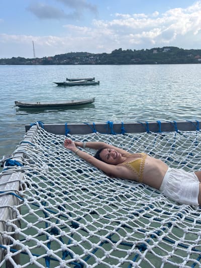A woman with dark hair is lying on a white net hammock suspended over water. She is wearing a yellow crocheted bikini top and white linen shorts. Her arms are stretched above her head, and she is smiling at the camera. The hammock is attached to wooden beams. In the background, there are several small boats on the water. Beyond the water, there is a coastline with trees and buildings, including a prominent telecommunications tower. The sky is partly cloudy. This scene is likely located in Nusa Penida, Indonesia, an island known for its scenic coastlines and ocean activities.
