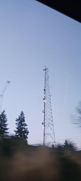 A tall, grey lattice-structured communications tower stands prominently against a light blue, largely clear sky. Multiple white and light-colored antennas, including several circular dish antennas and rectangular panel antennas, are affixed at various elevations along the tower's framework. To the left and slightly behind the tower, several dark green conifer trees are visible, their foliage appearing dense. The immediate foreground exhibits significant motion blur, indicating the image was captured from a moving vehicle. This blurred area consists of indistinct dark green and brownish hues, suggesting ground cover or other vegetation. A partial, dark, diagonal line is visible in the upper right, consistent with the interior edge of a vehicle's roofline or window frame. No people or specific human activity are visible. The lighting suggests ambient daylight, possibly early morning or late afternoon. The image provides no specific identifiable landmarks to confirm its location within Vancouver, United States.