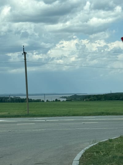 The image shows a landscape with a road on the right side, bordered by green grass. Adjacent to the grass is a wide, green field, possibly a pasture or farmland. Further in the background, a body of water is visible, with a line of trees running along its shore. On the left side of the image, the sky is cloudy with scattered power lines and a utility pole. This scene could be located in the city of Kryukovo, Russia, based on the landscape.