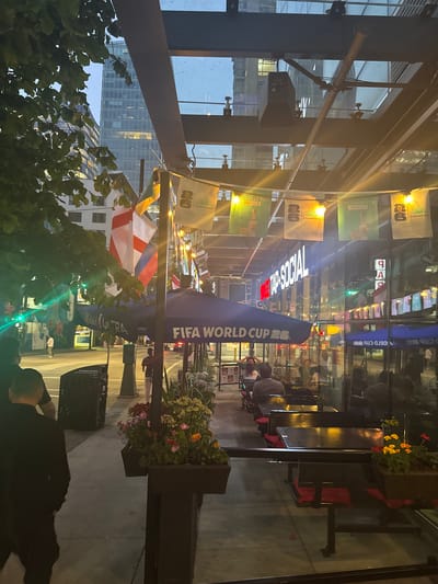 The image captures an outdoor patio area of a establishment called "TAP SOCIAL" located in Vancouver, Canada. The scene is viewed from street level, looking towards the building entrance and across the patio. 

In the foreground, the edge of a dark-colored planter with colorful flowers is visible on the right. Several tables with black surfaces and red seating are arranged on the patio, some occupied by patrons. A large blue umbrella with "FIFA WORLD CUP 26" printed on it is positioned over some of the tables. 

Hanging above the patio is a transparent roof structure supported by dark beams. Strung across the beams are banners, some featuring national flags and others promoting "CASAMIGOS" and the "FIFA WORLD CUP". String lights illuminate the area, casting a warm glow on the scene.

In the background, the exterior of the "TAP SOCIAL" establishment is visible, with large glass windows reflecting the street and surrounding buildings. A street with cars and pedestrians is visible beyond the patio. Tall buildings, characteristic of a city skyline, are visible in the distance. 

Several individuals are present in the image, some seated at tables on the patio and others walking on the sidewalk or street. The overall atmosphere suggests an evening setting, possibly during a sporting event or a social gathering.