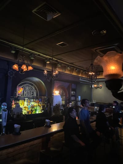 The image depicts the interior of a dimly lit bar with a prominent bar area filled with an assortment of liquor bottles. A large archway signage above the bar reads "CAMDEN." Several individuals are visible, some standing near the bar and others seated in front of it, appearing to be patrons. Decorative lighting fixtures, including chandeliers and lamps, are present throughout the space, contributing to the ambient mood. The overall environment suggests a social gathering or a night out at a bar. The location is identified as Central Jakarta, Indonesia.
