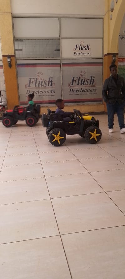 The image displays an indoor commercial area, likely a shopping mall corridor, located in Ngong ward, Kenya. The floor is composed of large, light-colored square tiles. The background features a storefront with multiple glass panels, prominently displaying "Flush Drycleaners" branding, including a hanger logo and the slogan "DRYCLEANERS OF DISTINCTION & EXCELLENCE." Structural columns with a textured yellowish-brown finish are visible; one on the left contains a dark sconce-style light fixture, and another on the right exhibits decorative arch cutouts.
Two young children, estimated to be between 3-5 years old, are operating electric ride-on toy vehicles. In the foreground, a child wearing dark clothing is seated in a black ride-on car with yellow accents and yellow star-spoked wheels, gripping the steering wheel and facing towards the right side of the frame. Further back on the left, another child, wearing a green top, is seated in a red and black ride-on car, positioned with their back to the camera. An adult male stands upright to the right, in the background, observing the scene. He is dressed in a dark jacket, blue jeans, and white sneakers, with his hands placed in his pockets.
