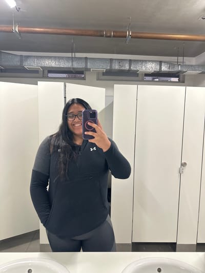 A person is taking a selfie in a public bathroom with white stalls and a sink, potentially located in Santiago, Chile. The person, wearing glasses and athletic attire, holds a smartphone up to capture the image. The background reveals standard bathroom fixtures such as pipes and ventilation shafts.