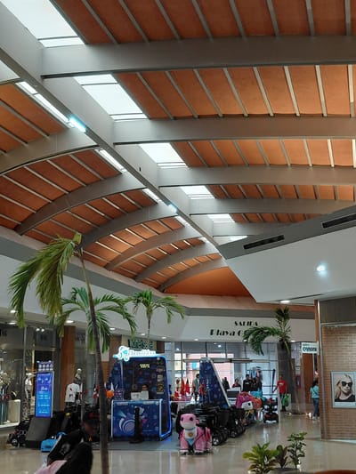 The image depicts the interior of a shopping mall, likely in Pampatar, Venezuela, featuring an arched ceiling with translucent panels and orange fabric. Palm trees are visible throughout the space. In the foreground, a child's ride is parked, along with a kiosk offering a game. People are seen browsing shops and walking through the mall. Signage includes "SALIDA" (Exit) and "Playa G" for a store. To the right, there's an optical shop called "CARONI" and a framed portrait of a woman wearing sunglasses. The environment appears to be well-lit and spacious, typical of a modern shopping center.