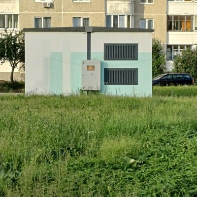 This image depicts an outdoor urban scene in Baranavichy, Belarus, dominated by a rectangular, single-story utility building positioned in the midground. The building features an upper section painted white or light grey and a lower section painted light teal. A dark grey vertical conduit extends from the roofline down the center of the building to a grey electrical panel box mounted on the teal section. To the right of the conduit, two horizontal dark grey louvered vents are installed on the building's facade.
The foreground is covered with dense, unkempt green grass and various herbaceous plants. In the background, several multi-story residential buildings with textured, light-colored facades are visible. These buildings exhibit multiple windows, some with white frames, and at least one air conditioning unit is visible on a facade. A small deciduous tree with green foliage is situated to the left of the utility building. On the right side of the scene, a dark-colored station wagon is partially visible behind the utility building and a cluster of green bushes. The lighting suggests daytime conditions. There are no individuals present in the frame.