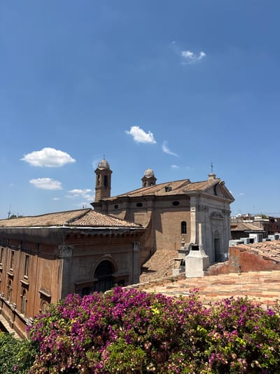 A high-angle, eye-level, medium, outdoor shot captures a view of Roman rooftops under a bright blue sky with scattered white clouds. In the foreground, a dense cluster of vibrant pink bougainvillea flowers spills over a rooftop, obscuring some of the lower architectural details. Behind the flowers, the terracotta-tiled roofs of several buildings are visible, some appearing aged and weathered.
Dominating the mid-ground is a large, brick building with a pale stone facade, likely a church or similar religious structure. It features a central dome with a cross on top, and a tall, separate bell tower with arched openings and a smaller dome. The building has several arched windows and decorative architectural elements.
Further in the background, more rooftops and buildings are visible, some with visible air conditioning units. In the far distance, a metal structure, possibly a crane or tower, pierces the skyline. The overall scene suggests a sunny day in a historic urban environment. The style of the architecture, particularly the bell tower and the church facade, strongly points to a location within Rome, Italy, likely in a historic district with characteristic tiled roofs.