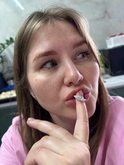 The image depicts a close-up of an adult female, situated in Gornyi (Dvurechenskoe S/P), Russia, captured from the chest upwards. She has light brown hair, blue eyes, and fair skin with a singular brown nevus on her right cheek, inferior to the lateral canthus. The subject wears a light pink garment. Her right index finger, displaying a pale pink manicured nail, is positioned against her lips. A ring, featuring five clear, circular faceted stones set into a silver-toned band, rests on the fingertip, positioned at the philtrum area. Her gaze is directed upwards and to the right of the frame. The background is out of focus, showing blurred elements including a linear light fixture and green foliage on the left, and a white plastic storage basket on the right.
