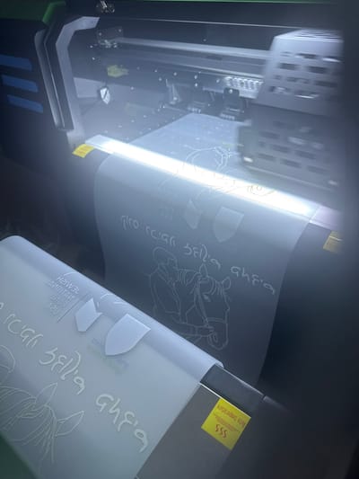 An industrial-grade, large-format printer is depicted in operation within a facility located in Rishon LeZion, Israel. The machine is actively processing a continuous roll of light-colored material, likely a film or vinyl, which is being fed through the printing mechanism. A prominent internal light source illuminates the area where designs are being applied. The material exiting the printer displays printed graphics in white or light gray outline against its dark substrate. These graphics include a repeating pattern featuring a person with a horse, Hebrew script, and the English text "JEWISH NATIONAL FUND USA," along with various geometric or shield-like emblems. A yellow sticker bearing the text "Note temperature" and "SSS" is visible on the printer's lower right section. No individuals are present in the scene.