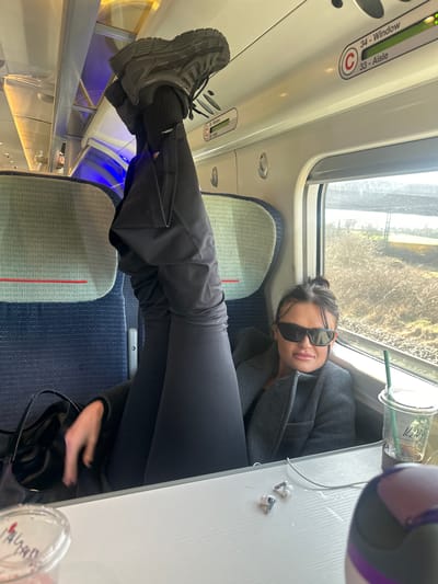 A person is observed seated in a window seat aboard a modern passenger train, operating in the vicinity of Stafford, United Kingdom. The individual, an adult female with dark hair styled in a bun, wears dark sunglasses, a grey coat, black trousers, black socks, and black athletic shoes. Her right leg is extended vertically, with the sole of her black shoe resting against the overhead luggage rack. Her left hand is positioned near a black shoulder bag. She maintains a direct gaze towards the camera. A white fold-down table is positioned in front of her, upon which rests a pair of white wireless earbuds, a clear plastic Starbucks cup containing an iced beverage with a green straw, and a partially visible clear plastic cup and a purple and grey bottle. The train's interior features blue and green patterned seating. Overhead signage displays "34 - Window" and "33 - Aisle" with a green illuminated indicator and a red 'C' symbol. Through the window, a sunlit exterior reveals dry, brown vegetation and apparent railway infrastructure.