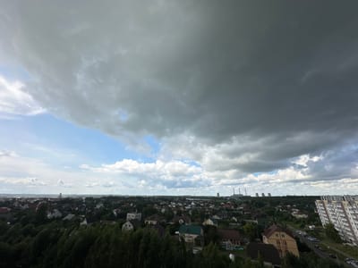 The image depicts an aerial view of a suburban landscape under a dramatic, overcast sky. Dark, heavy clouds dominate the upper portion of the frame, with a sliver of bright blue sky visible on the left, peeking through breaks in the cloud cover. A town, identified as being in the Shchomyslitsky Sel'skiy Soviet, Belarus, spreads across the middle ground, characterized by numerous houses with red, brown, and white roofs, interspersed with dense green trees. In the distance, beyond the town, industrial structures with tall smokestacks are visible on the horizon, suggesting a power plant or similar facility. To the far right, a modern, multi-story apartment building stands prominently, its white facade contrasting with the more traditional architecture of the town. The ground below is covered with lush green foliage, indicating a temperate climate. No people or specific actions are discernible in the image.